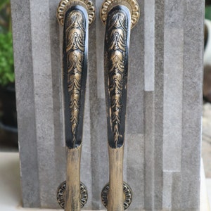 May include: Two ornate door handles with a black and gold finish. The handles have a detailed, textured design and are mounted on a gray stone surface. The handles are approximately 10 inches in length, with a decorative circular base.