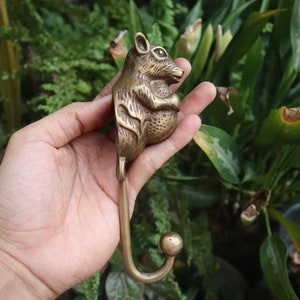 May include: A brass hook shaped like a squirrel, with a rounded head and a curled tail. The hook is held in a hand, with the background blurred.