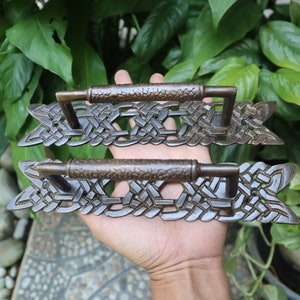 May include: Two ornate bronze door handles with intricate knotwork designs. The handles are held in a hand against a green leafy background.