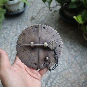 May include: A bronze-colored, round cabinet latch with a sunburst design. The latch features a central locking mechanism with a decorative key-shaped element and a small chain. The design is symmetrical and detailed, suggesting a vintage or antique style.