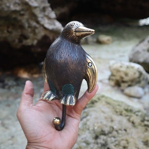 Puede incluir: Un gancho de pingüino de bronce con pico y patas doradas. El gancho tiene forma de pingüino con un diseño verde y dorado en el vientre.