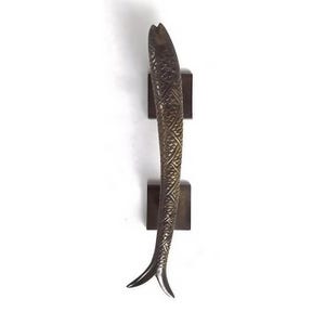 May include: Bronze-coloured door pull in the shape of a fish. The fish has a textured, scale-like pattern and is mounted on two square, dark brown blocks. The fish's tail is split into two fins. The door pull is mounted on a white background.