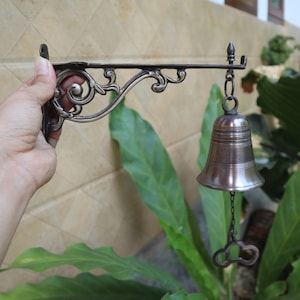 Può includere: Una campana in metallo argentato con una staffa decorata e ornata. La campana è appesa a una catena e ha un piccolo pezzo decorativo in cima.