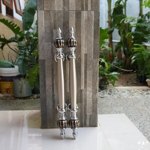 May include: Two silver door handles with decorative finials and center accents. The handles are vertically aligned and feature a combination of silver, gold, and black elements. The handles are mounted on a gray tiled surface.