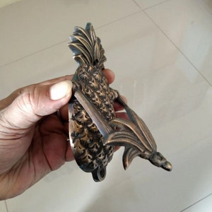 Puede incluir: Un llamador de puerta de pi&ntilde;a de lat&oacute;n con un dise&ntilde;o detallado. El llamador tiene forma de pi&ntilde;a con hojas y un anillo para llamar.