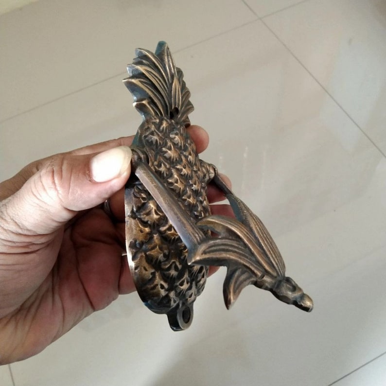 Puede incluir: Un llamador de puerta de pi&ntilde;a de lat&oacute;n con un dise&ntilde;o detallado. El llamador tiene forma de pi&ntilde;a con hojas y un anillo para llamar.