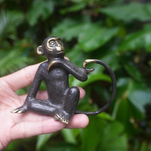 May include: A small, bronze-colored monkey figurine. The monkey is in a seated position with its tail curled. The details include gold-colored accents on the ears, hands, and feet. The background is a blurred view of green foliage.