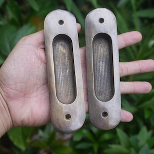 Pode incluir: Duas puxadores de porta de latão antigo com forma oval e um recorte retangular no centro. Os puxadores têm um acabamento desgastado e envelhecido.