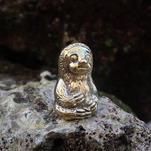 May include: A small, gold-colored sloth figurine sitting on a rock. The sloth is in a sitting position with its arms crossed in front of it.