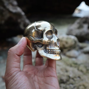 May include: A detailed brass skull sculpture with visible teeth and eye sockets. The skull is held in a hand, showcasing its intricate design and metallic sheen. The skull is a decorative item.