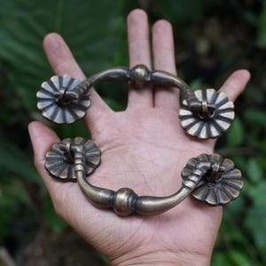 Puede incluir: Dos tiradores de cajón de latón de estilo antiguo con un diseño de asa curva y un detalle central redondeado. Cada extremo del asa está unido a una placa circular en forma de flor con ranuras radiales y un acabado oscuro.
