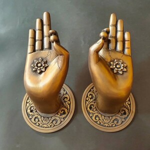 May include: Two ornate brass door handles shaped like hands in a gesture of prayer or blessing. Each handle features intricate floral designs and a raised knob on the index finger.