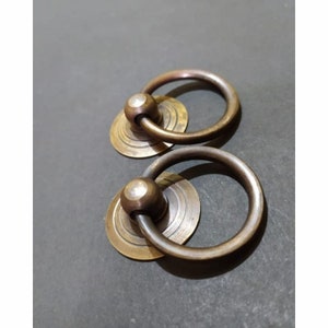 Pode incluir: Dois puxadores de anel de latão antigo com base circular. Os puxadores de anel têm um acabamento liso e envelhecido e são perfeitos para adicionar um toque de charme vintage a qualquer projeto.