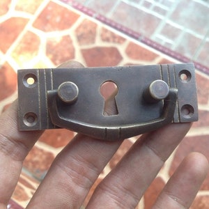 May include: Brass cabinet pull with a keyhole and a curved handle. The pull has a dark brown finish and is mounted on a rectangular plate.