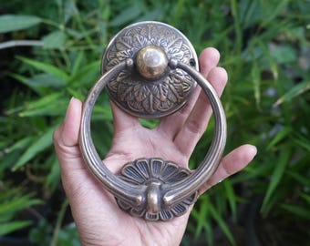 May include: Antique brass door knocker with a circular backplate and a ring handle. The backplate features an embossed floral design, and the handle is attached to a decorative base. The knocker is held against a backdrop of green foliage.