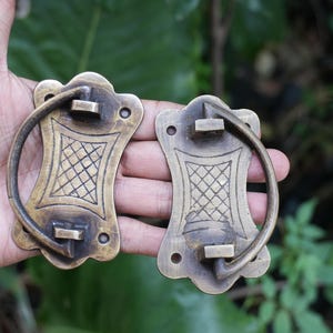 May include: Two antique-style brass drawer pulls featuring a rectangular backplate with scalloped edges and a central diamond-patterned engraving. Each pull includes a hinged, semi-circular handle attached to the backplate with two mounting holes.