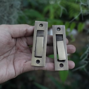 May include: Two rectangular brass flush pulls with a brushed metallic finish. Each pull features a central recessed lever with horizontal ribbed detailing at the top and two circular screw holes at the top and bottom for mounting purposes.