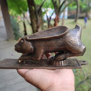 May include: A brass door knocker shaped like a rabbit with its ears raised. The rabbit is in a crouching position with its front paws extended. The knocker is mounted on a rectangular brass plate.