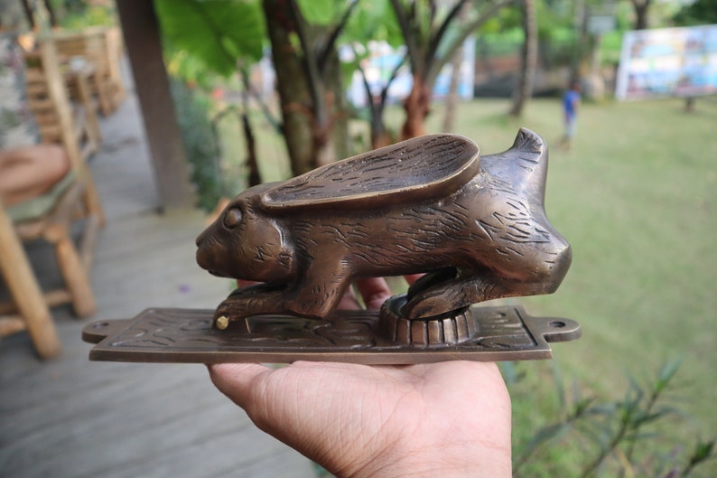 May include: A brass door knocker shaped like a rabbit with its ears raised. The rabbit is in a crouching position with its front paws extended. The knocker is mounted on a rectangular brass plate.