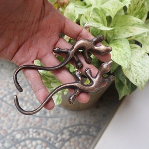 Könnte beinhalten: Zwei bronzefarbene Gecko-Haken mit einem eingerollten Schwanz. Die Haken sind in Form eines Geckos mit ausgestreckten Beinen. Die Haken werden in einer Hand gehalten.