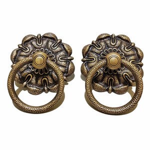 May include: Two antique brass cabinet pulls with a floral design and a ring handle. The pulls have a textured finish and a distressed look.