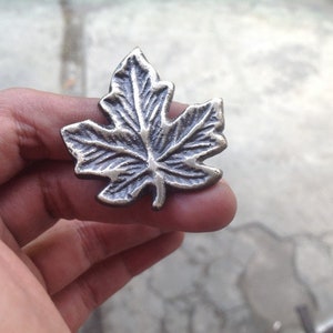 May include: A silver-toned metal maple leaf charm. The leaf is detailed with veins and has a slightly textured surface.
