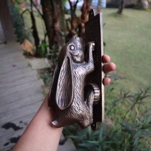 May include: A bronze rabbit door knocker with a detailed design. The rabbit is holding a door handle and is mounted on a rectangular plaque.