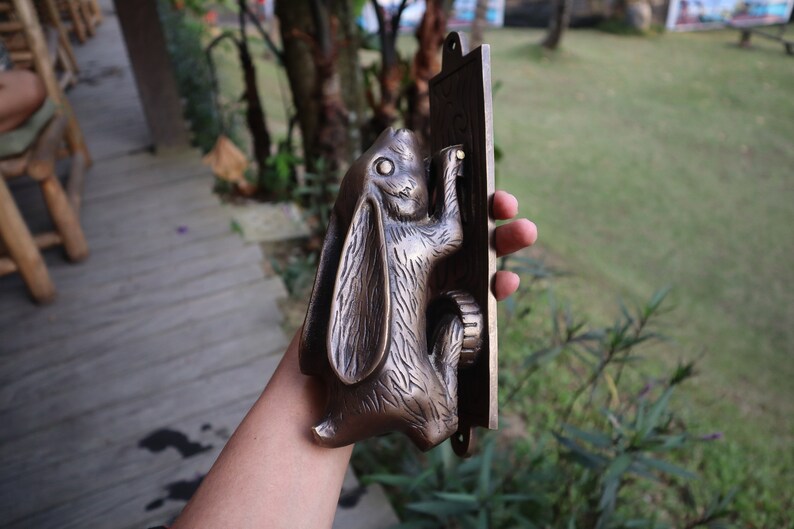 May include: A bronze rabbit door knocker with a detailed design. The rabbit is holding a door handle and is mounted on a rectangular plaque.