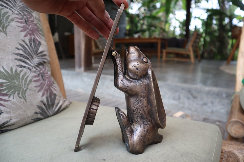 May include: A bronze rabbit door knocker with a raised paw and a detailed design. The knocker is attached to a rectangular plate with a decorative edge.