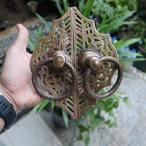 Könnte beinhalten: Verzierter Türgriff aus Messing mit einem dekorativen, filigranen Design. Der Griff verfügt über zwei runde Ringe mit einem detaillierten Blumenmuster.