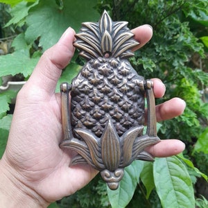Puede incluir: Un llamador de puerta de pi&ntilde;a de lat&oacute;n con un dise&ntilde;o detallado. El llamador tiene la forma de una pi&ntilde;a con hojas y una superficie texturizada.