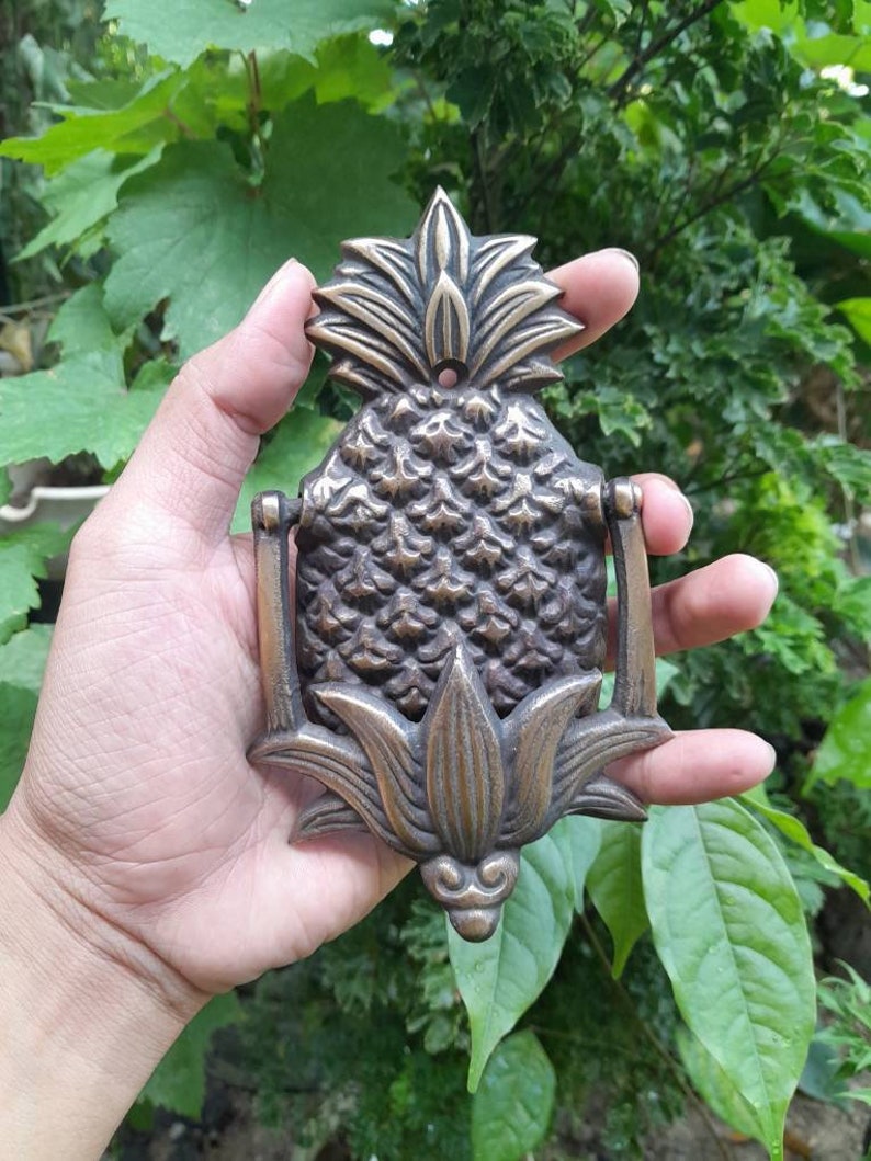 Puede incluir: Un llamador de puerta de pi&ntilde;a de lat&oacute;n con un dise&ntilde;o detallado. El llamador tiene la forma de una pi&ntilde;a con hojas y una superficie texturizada.