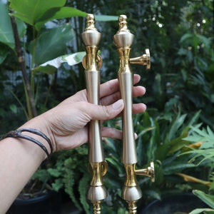 May include: A pair of gold door handles with a decorative design. The handles are held in a hand with a bracelet.