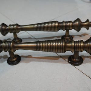 Könnte beinhalten: Zwei antike Türgriffe mit verzierten, detaillierten Designs. Die Griffe sind aus Metall und haben eine dunkelbraune Bronze-Oberfläche. Sie verfügen über eine gerippte Textur und dekorative Endstücke.