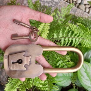May include: A brass padlock with a decorative design and two keys. The padlock has a U-shaped shackle and a keyhole with a small, decorative knob. The keys are both made of brass and have a simple design.