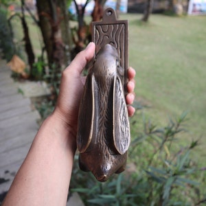 May include: A bronze rabbit door knocker with a detailed design. The knocker is held in a hand and the background is blurred.