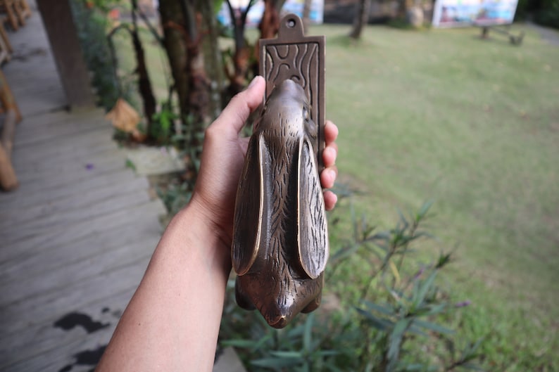 May include: A bronze rabbit door knocker with a detailed design. The knocker is held in a hand and the background is blurred.
