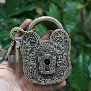 May include: An antique brass padlock with two keys. The lock is decorated with detailed floral and scroll patterns. The keys are attached to a ring. The background is a blurred view of green leaves.