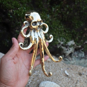 Puede incluir: Un gancho de metal dorado en forma de cráneo de pulpo con cinco tentáculos. El gancho está diseñado para colgar en una pared.