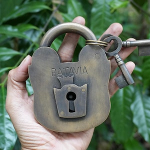 May include: An antique brass padlock with the word "BATAVIA" engraved on the front. Two matching keys are attached to the padlock. The padlock is held in front of a background of green leaves.