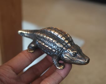 May include: A small, detailed bronze alligator figurine with a textured, black and gold finish. The alligator is posed with its mouth closed and its head turned slightly to the side.