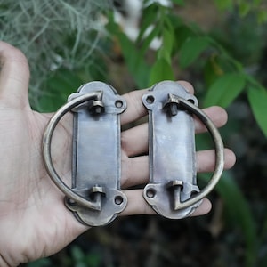 May include: Two antique-style brass drawer pulls with a rectangular backplate and a semi-circular swinging handle. Each backplate features rounded ends with two screw holes for mounting, showcasing a weathered, dark metallic finish.
