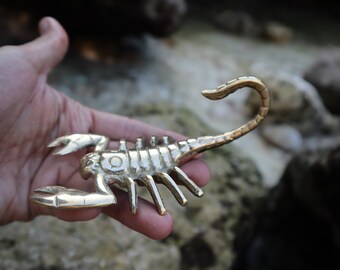 May include: A gold-colored metal scorpion figurine with detailed features. The scorpion is posed with its tail curled up and its claws extended.