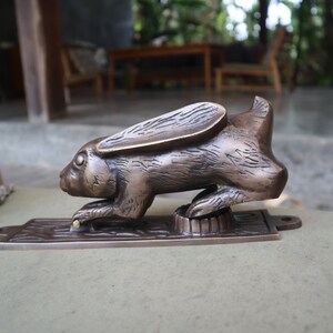 May include: A bronze rabbit door knocker with a detailed design. The rabbit is in a crouching position with its ears raised. The knocker is mounted on a rectangular base.