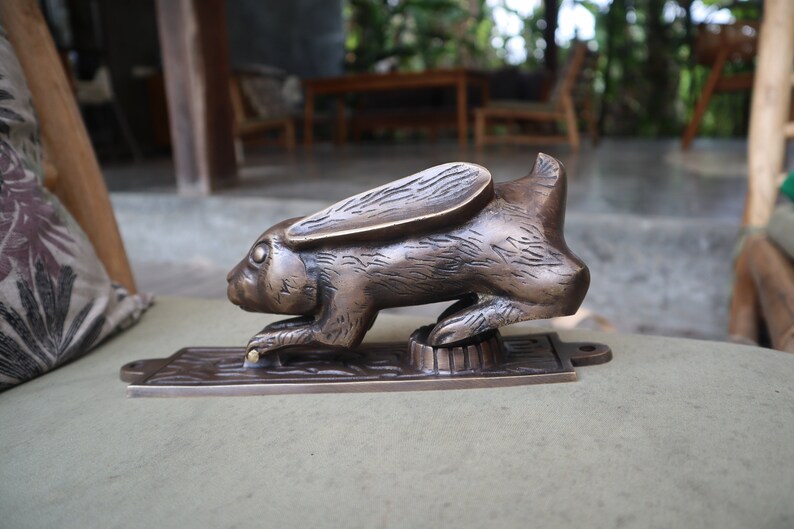 May include: A bronze rabbit door knocker with a detailed design. The rabbit is in a crouching position with its ears raised. The knocker is mounted on a rectangular base.