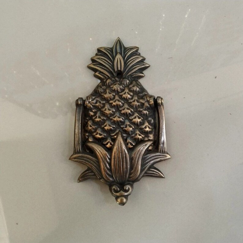 Puede incluir: Un llamador de puerta de pi&ntilde;a de lat&oacute;n con un dise&ntilde;o detallado. El llamador tiene la forma de una pi&ntilde;a con una parte superior de hojas y un anillo para llamar.