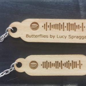 Peut inclure: Deux porte-clés en bois avec un logo Spotify et un code-barres. Le porte-clés du haut porte le texte "Butterflies by Lucy Spraggan".