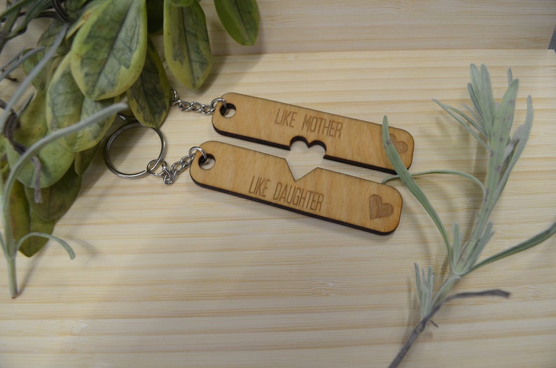 May include: Two wooden keychains with a heart-shaped cutout. The top keychain says "Like Mother" and the bottom keychain says "Like Daughter".