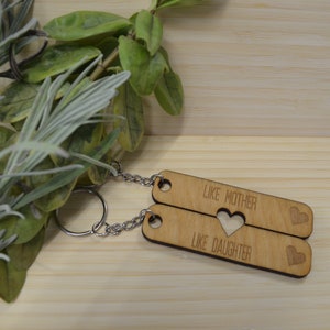 May include: Two wooden keychains with the text "Like Mother Like Daughter" and a heart cut-out in the middle. The keychains are connected by a silver chain.