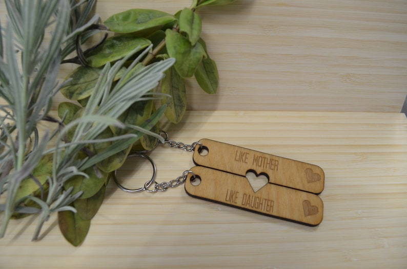 May include: Two wooden keychains with the text "Like Mother Like Daughter" and a heart cut-out in the middle. The keychains are connected by a silver chain.